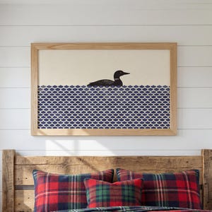 May include: A bedroom scene featuring a wooden headboard and a bed covered with a red, green, and navy plaid comforter and pillows. Above the bed is a framed print of a loon on blue water. A rustic lantern sits on a wooden side table.