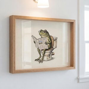 Könnte beinhalten: Gerahmter Kunstdruck mit einem grünen Frosch mit Brille, der die Zeitung "The Daily Croak" auf einer Toilette liest. Das Kunstwerk ist in einem hellbraunen Holzrahmen eingefasst.