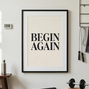 Può includere: Una stampa incorniciata con le parole "BEGIN AGAIN" in grassetto nero su sfondo color crema. La cornice è nera. Un orologio digitale, una bottiglia d'acqua e attrezzature per l'allenamento sono visibili sullo sfondo.
