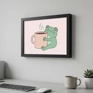 Puede incluir: Una impresión enmarcada con una rana de dibujos animados con ojos somnolientos, abrazando una taza de café beige. La rana es de color verde claro y la taza tiene vapor saliendo. El fondo es de color rosa suave.