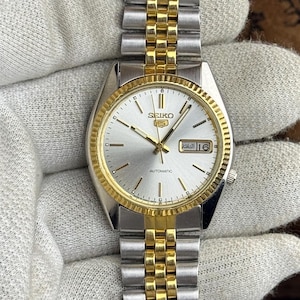 May include: A Seiko 5 automatic wristwatch featuring a silver dial with gold-tone hour markers and hands. The watch has a fluted gold-tone bezel and a two-tone stainless steel jubilee-style bracelet with alternating silver and gold-tone links.