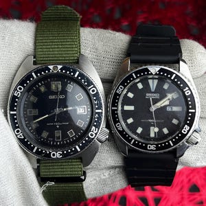 May include: Two vintage Seiko dive watches. One has a green nylon strap, the other a black rubber strap. Both watches have black dials with luminous markers and hands, and rotating bezels with minute markings. The watch faces display the Seiko logo and other text.