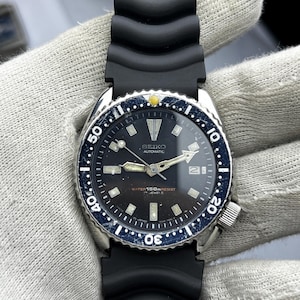 May include: A vintage Seiko Automatic wristwatch with a black dial, luminous hands, and a date window. The watch features a rotating blue bezel, a black rubber strap, and the text "WATER 150M RESIST".