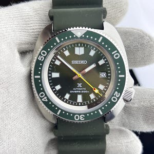 Può includere: Orologio subacqueo automatico Seiko con quadrante e lunetta verdi. L'orologio presenta una cassa in acciaio inossidabile, un cinturino in gomma verde e un datario a ore 3. Impermeabile fino a 200 metri.