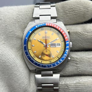 Op de afbeelding: Een vintage Seiko chronograaf horloge met een goudkleurige wijzerplaat, met een blauwe en rode tachymeter bezel. Het horloge heeft een roestvrijstalen band en een datumweergave op 3 uur. De wijzerplaat toont de tekst "SEIKO CHRONOGRAPH AUTOMATIC".