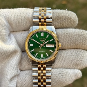 May include: A Seiko 5 automatic wristwatch featuring a vibrant green dial, silver and gold-tone fluted bezel, and a matching two-tone stainless steel link bracelet. The dial displays the Seiko logo, day-date window, and luminous hour markers.