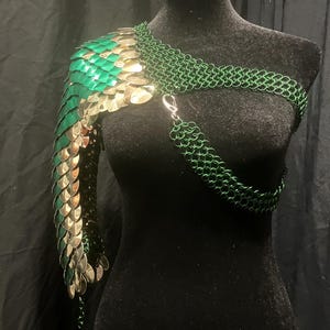 Puede incluir: Una pieza de armadura de cota de malla para un solo hombro con una manga construida con escamas metálicas verdes y plateadas superpuestas. La sección superior y la correa del pecho están hechas de anillos de metal verde oscuro entrelazados.