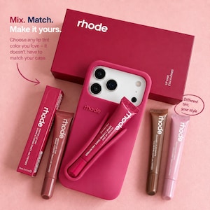 Puede incluir: Una funda de silicona para teléfono de color magenta de la marca rhode con un soporte integrado para un bálsamo labial, acompañada de una caja magenta a juego y tres tubos de bálsamo labial rhode en tonos marrón y rosa, con texto impreso.