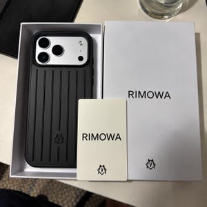 Puede incluir: Una funda de smartphone negra con textura de ranuras verticales y un módulo de cámara plateado, presentada en una caja blanca junto a una tarjeta blanca. Tanto la caja como la tarjeta muestran el texto negro RIMOWA y un pequeño logotipo negro de una M estilizada.