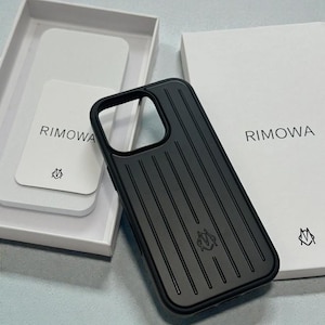Op de afbeelding: Zwarte telefoonhoes met verticale groeven en een logo. De hoes ligt naast een witte doos en een kaart, beide met de merknaam "RIMOWA" erop gedrukt. De hoes heeft een strak, modern design.