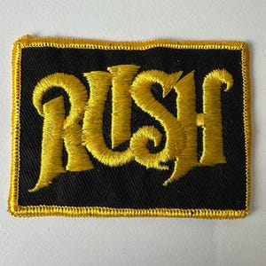 May include: A rectangular embroidered patch featuring the word RUSH in bold, yellow, stylized lettering centered on a black fabric background. The entire perimeter of the patch is finished with a thick, yellow stitched border.
