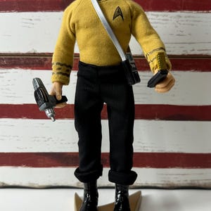 May include: A Star Trek action figure wearing a mustard yellow long-sleeve shirt with a black collar and black trousers. The figure holds a gray phaser in the right hand and a communicator in the left, with a silver shoulder strap and black boots.