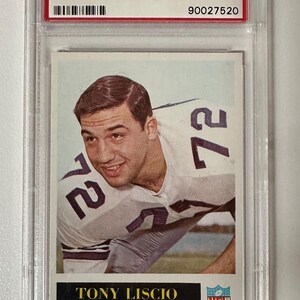 May include: A 1965 Philadelphia Tony Liscio football card, graded NM-MT 8 by PSA. The card features a color image of Tony Liscio in a Dallas Cowboys uniform, with the number 72 visible. The card is encased in a clear protective holder.
