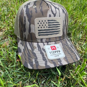 May include: A camouflage baseball cap with a mesh back and a curved brim. The front features a leather patch with a distressed American flag design. The cap has the text "112PFP PRINTED 5 PANEL OSFM" on the brim.