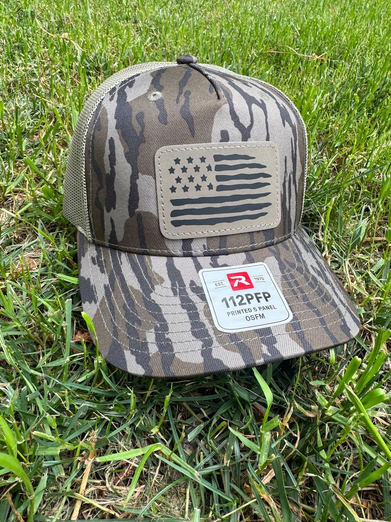 May include: A camouflage baseball cap with a mesh back and a curved brim. The front features a leather patch with a distressed American flag design. The cap has the text "112PFP PRINTED 5 PANEL OSFM" on the brim.
