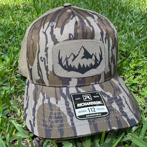 May include: Camouflage-patterned trucker hat with a mountain and tree patch. The hat has a mesh back and a curved brim. The patch features a black mountain range silhouette. The hat is adjustable and has the Richardson brand label.