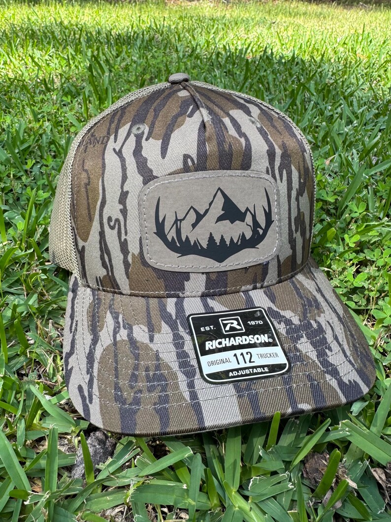 May include: Camouflage-patterned trucker hat with a mountain and tree patch. The hat has a mesh back and a curved brim. The patch features a black mountain range silhouette. The hat is adjustable and has the Richardson brand label.