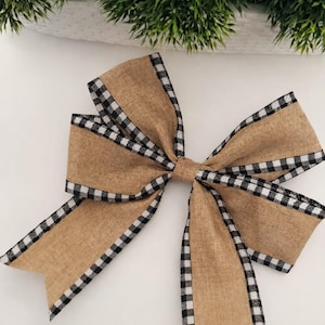 May include: A large decorative bow with a tan burlap center and black and white checkered trim. The bow is centered and has multiple loops and tails. Greenery is visible in the background.