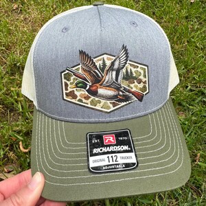 May include: Gray and olive green Richardson 112 Trucker hat with a duck graphic. The hat features a gray crown, a white mesh back, and an olive green brim. The duck graphic is in a camouflage hexagon.