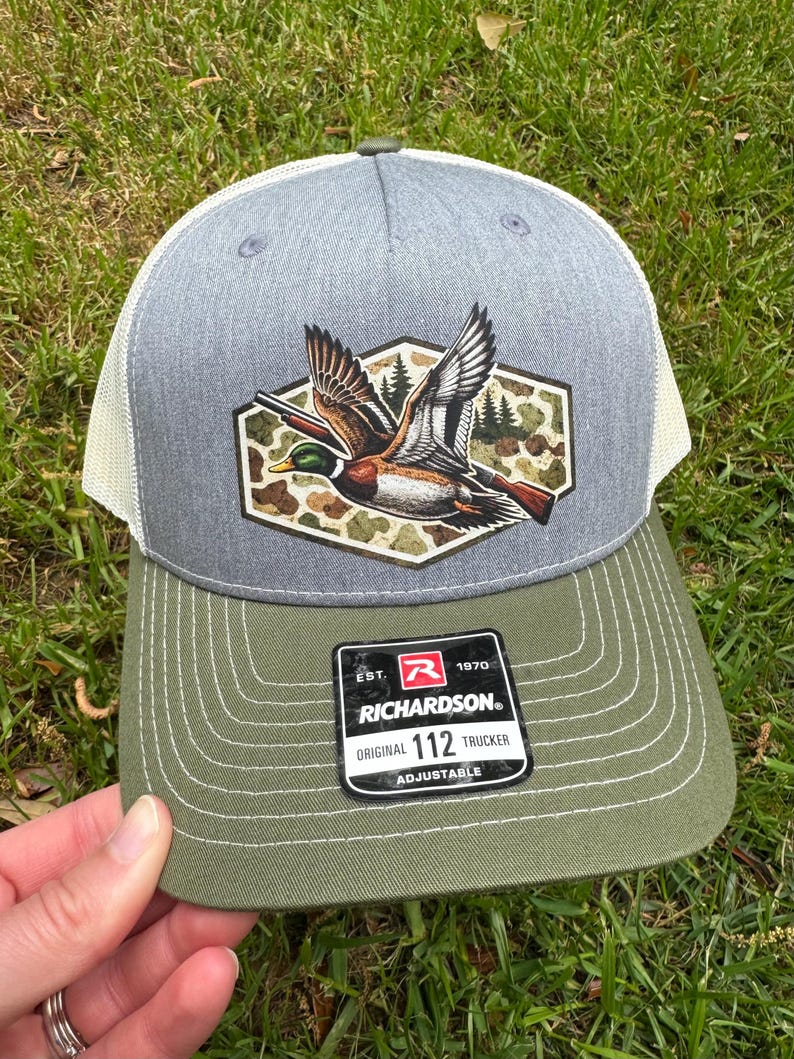 May include: Gray and olive green Richardson 112 Trucker hat with a duck graphic. The hat features a gray crown, a white mesh back, and an olive green brim. The duck graphic is in a camouflage hexagon.