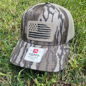 May include: A camouflage-patterned baseball cap with a mesh back and a curved brim. The cap features a patch with a black and tan American flag design. The tag reads "112PFP PRINTED 5 PANEL OSFM".