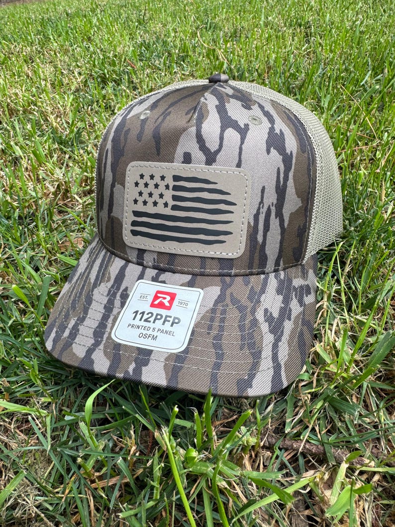 May include: A camouflage-patterned baseball cap with a mesh back and a curved brim. The cap features a patch with a black and tan American flag design. The tag reads "112PFP PRINTED 5 PANEL OSFM".