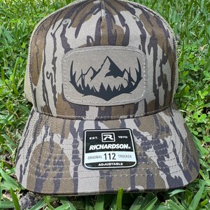 May include: A camouflage-patterned Richardson 112 Trucker hat with a mountain and forest patch. The hat features a brown and tan color scheme, a mesh back, and an adjustable strap. The patch is a light brown color with a black mountain and forest design.