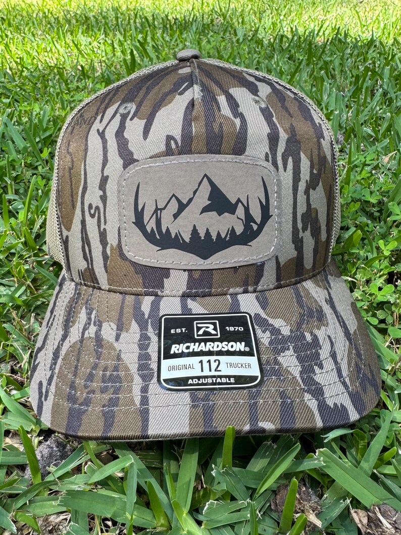 May include: A camouflage-patterned Richardson 112 Trucker hat with a mountain and forest patch. The hat features a brown and tan color scheme, a mesh back, and an adjustable strap. The patch is a light brown color with a black mountain and forest design.
