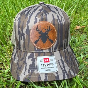 May include: A camouflage-patterned baseball cap with a brown leather patch featuring a deer silhouette and crossed rifles. The cap has a mesh back and a tag that reads "112PFP PRINTED 5 PANEL OSFM".