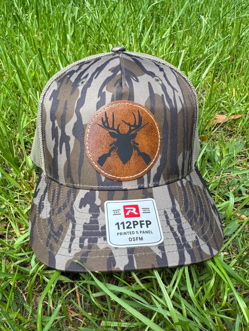May include: A camouflage-patterned baseball cap with a brown leather patch featuring a deer silhouette and crossed rifles. The cap has a mesh back and a tag that reads "112PFP PRINTED 5 PANEL OSFM".