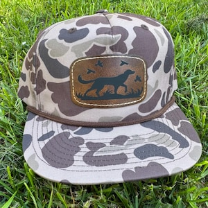 May include: Camouflage baseball cap with a brown leather patch featuring a dog silhouette, grass, and flying birds. The cap has a brown rope detail and is set against a background of green grass.