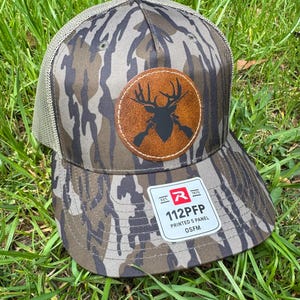 May include: A camouflage-patterned baseball cap with a brown leather patch featuring a black deer silhouette and crossed rifles. The cap has a mesh back and a curved brim. The tag reads "112PFP PRINTED 5 PANEL OSFM".