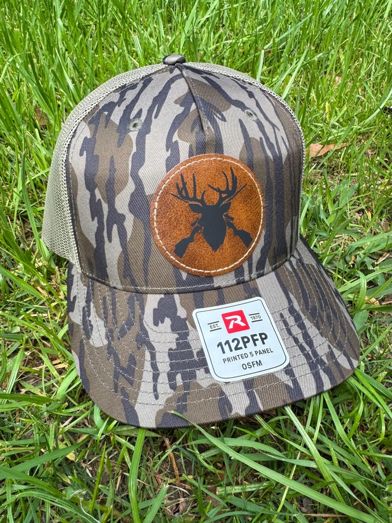 May include: A camouflage-patterned baseball cap with a brown leather patch featuring a black deer silhouette and crossed rifles. The cap has a mesh back and a curved brim. The tag reads "112PFP PRINTED 5 PANEL OSFM".