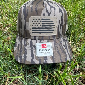 May include: Camouflage baseball cap with a mesh back and a leather patch featuring a distressed American flag design. The cap has a curved brim and a tag that reads "112PFP PRINTED 5 PANEL OSFM".