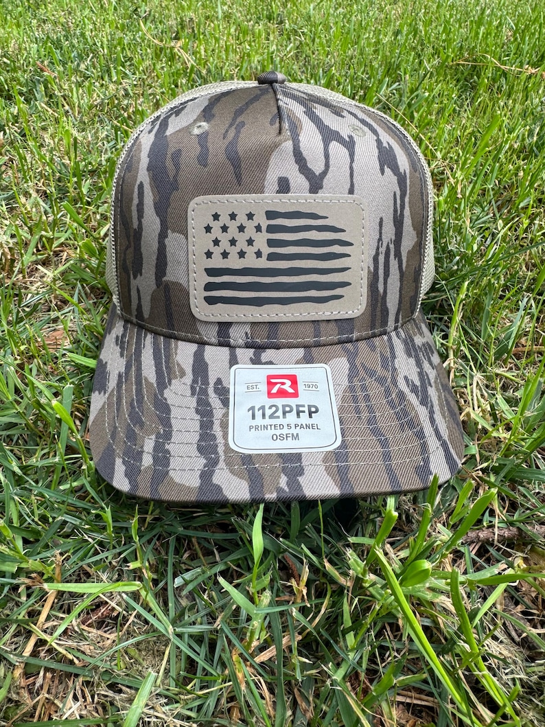 May include: Camouflage baseball cap with a mesh back and a leather patch featuring a distressed American flag design. The cap has a curved brim and a tag that reads "112PFP PRINTED 5 PANEL OSFM".