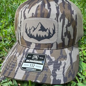 May include: A camouflage trucker hat with a mountain and forest patch. The hat features a brown and green camouflage pattern, a mesh back, and a curved brim. The patch has a black mountain and tree design. The tag reads "RICHARDSON ORIGINAL 112 TRUCKER ADJUSTABLE."