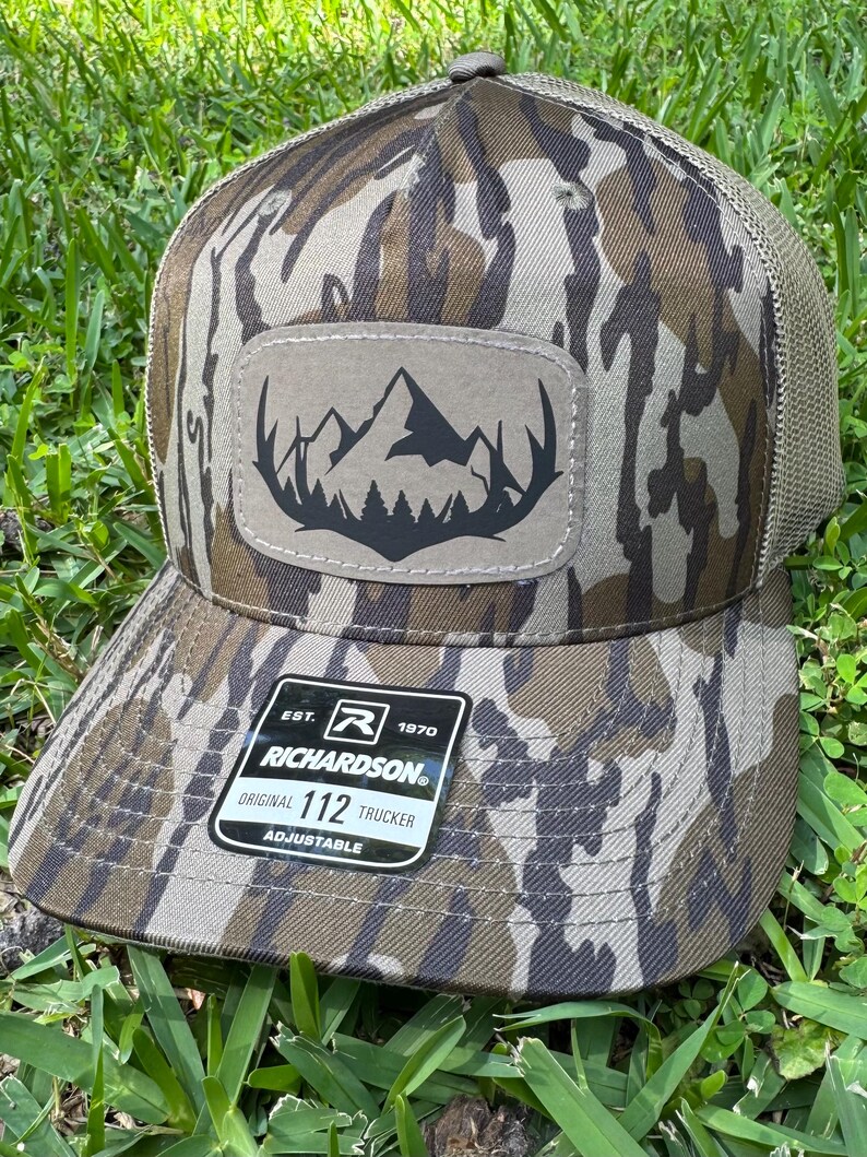 May include: A camouflage trucker hat with a mountain and forest patch. The hat features a brown and green camouflage pattern, a mesh back, and a curved brim. The patch has a black mountain and tree design. The tag reads "RICHARDSON ORIGINAL 112 TRUCKER ADJUSTABLE."