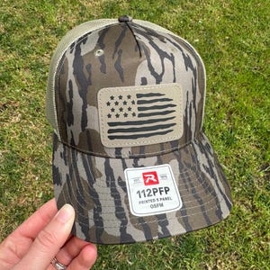 May include: A camouflage-patterned baseball cap with a mesh back and a patch featuring a stylized American flag. The cap has the text "112PFP PRINTED 5 PANEL OSFM" on a tag.