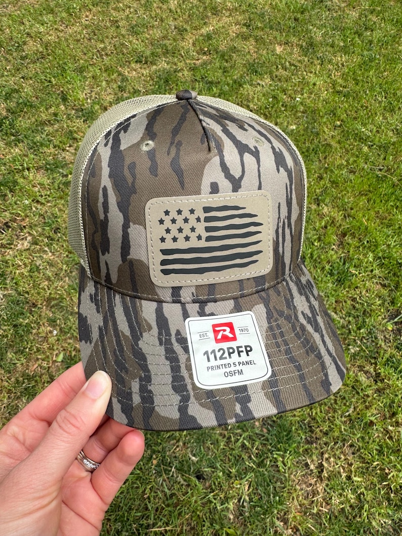 May include: A camouflage-patterned baseball cap with a mesh back and a patch featuring a stylized American flag. The cap has the text "112PFP PRINTED 5 PANEL OSFM" on a tag.