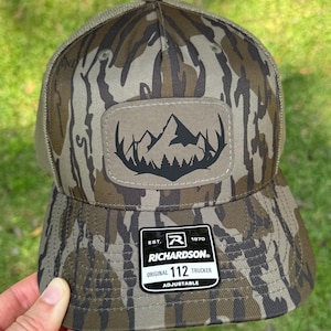 May include: A camouflage trucker hat with a mountain and tree patch. The hat has a curved brim and mesh back. The patch is a light brown color with a black mountain and tree design. The hat is adjustable and has a Richardson tag.