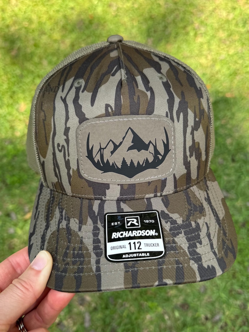 May include: A camouflage trucker hat with a mountain and tree patch. The hat has a curved brim and mesh back. The patch is a light brown color with a black mountain and tree design. The hat is adjustable and has a Richardson tag.