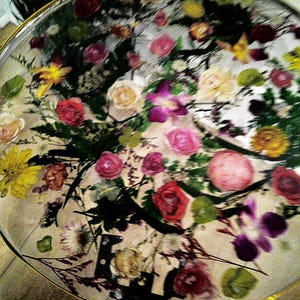 May include: A round, clear table top with a gold rim, filled with an assortment of colorful dried flowers. The arrangement includes red, pink, yellow, and purple blooms, with green foliage. The table top is on a light-colored wooden surface.