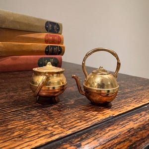 May include: A miniature gold-coloured tea set, comprising a small teapot and a small pot with a lid. The set is displayed on a dark wooden surface, with a stack of books in the background.
