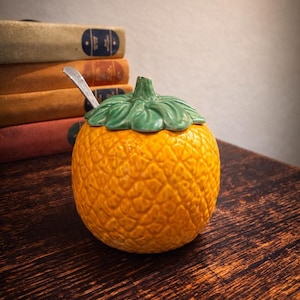 May include: A ceramic pineapple-shaped container with a green leafy lid and a small silver spoon. The container is a vibrant orange color and sits on a dark wooden surface. The background includes a stack of books.