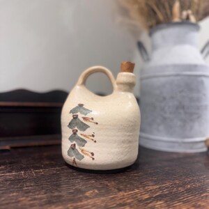 May include: A small, off-white ceramic jug with a handle and a cork stopper. The jug is decorated with stylized, hand-painted fish in green and brown. The jug sits on a wooden surface. The jug is approximately 15 cm tall.