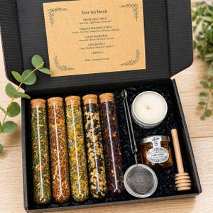 May include: A black gift box containing tea blends, a candle, honey, and a tea infuser. The tea blends are in glass tubes with cork stoppers. The box includes a card with tea blend names: Fresh Mint Apple, Golden Rooibos Citrus, Calm Chamomile, Ginger Spice, and Hibiscus.
