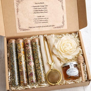May include: A gift box with tea blends, honey, and accessories. The box contains glass tubes filled with tea, a honey jar, a tea infuser, a wooden honey dipper, and a decorative flower. The text on the box reads "Your tea blends."