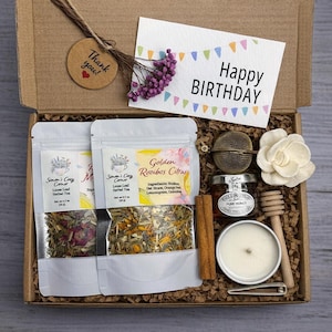 May include: A cardboard gift box containing two pouches of loose leaf herbal tea, a small jar of honey, a wooden honey dipper, a cinnamon stick, a metal tea infuser, a white candle, a white flower, and a card reading Happy BIRTHDAY.