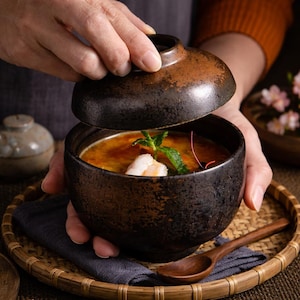 Puede incluir: Un cuenco de cerámica marrón oscuro con tapa, lleno de una sopa amarillo-naranja que contiene un camarón y adornado con una ramita de menta. El cuenco está sobre una bandeja tejida con una cuchara de madera y un paño azul oscuro.