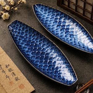 Può includere: Due piatti da portata in ceramica blu e bianca con un motivo a squame di pesce. I piatti ovali hanno un bordo dorato. Lo sfondo include un rametto di fiori bianchi e un pezzo di carta con caratteri asiatici.